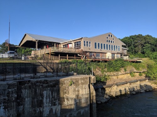 A large building with a deck overlooking a river, surrounded by greenery and a clear blue sky.