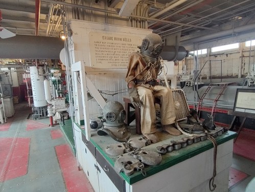 A vintage diving suit displayed on a platform in an industrial engine room, surrounded by various tools and equipment.