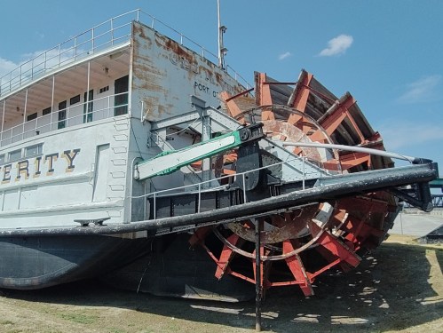 A large, weathered riverboat with a prominent paddlewheel, docked under a clear blue sky.
