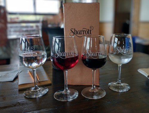 Four wine glasses with different types of wine, alongside a menu from Sharrott Winery, on a wooden table.