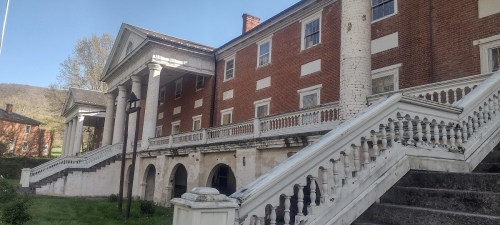 Historic brick building with white columns and a grand staircase, set against a backdrop of trees and hills.
