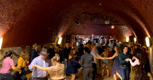 A lively dance floor in a dimly lit venue, with people dancing and a live band performing in the background.