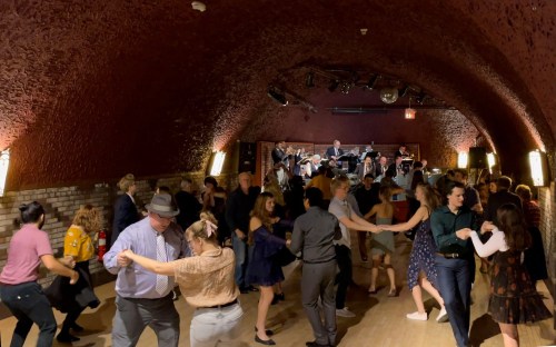 A lively dance floor with people dancing to a live band in a warmly lit, arched venue.