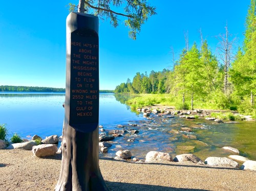 A tall wooden marker by a serene lake, detailing the Mississippi River's journey to the Gulf of Mexico.