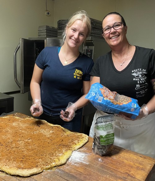 Two women are smiling while preparing dough topped with cinnamon and sugar in a kitchen. One holds a bag of ingredients.