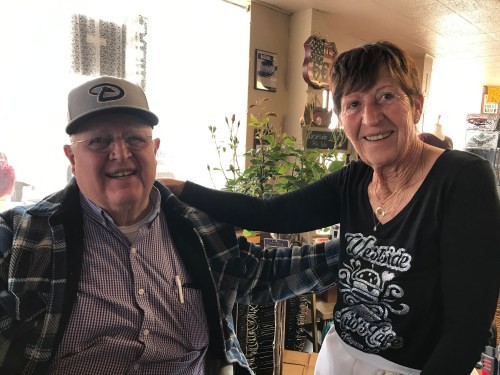 A smiling elderly man in a cap poses with a cheerful woman in a restaurant, both enjoying their time together.