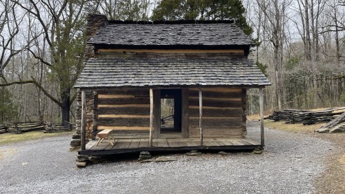 A rustic log cabin with a shingled roof, surrounded by bare trees and gravel pathways.