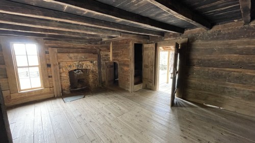 Interior of a rustic wooden room featuring a fireplace, wooden beams, and two doorways leading outside.