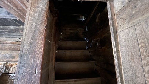 A dimly lit wooden staircase leading up, surrounded by rustic wooden walls with visible grain and texture.