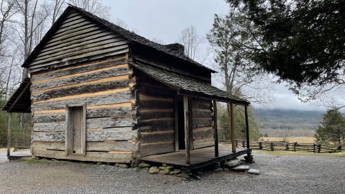 A rustic log cabin with a porch, surrounded by trees and a scenic landscape in the background.