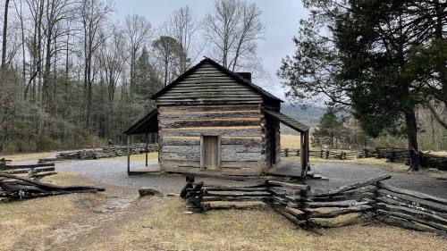 A rustic wooden cabin surrounded by a split-rail fence, set in a wooded area with bare trees and a cloudy sky.