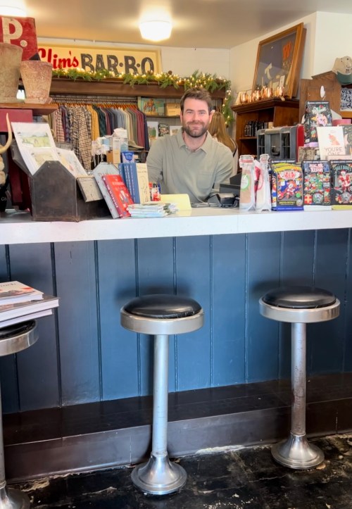 A smiling man stands behind a counter in a cozy shop, with stools in front and various items displayed around him.