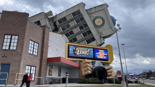 A tilted building with a large camera sculpture at the entrance, under a cloudy sky.