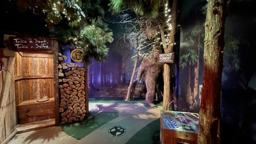 A whimsical indoor scene featuring a Bigfoot figure in a forest setting, with a selfie spot and wooden decor.