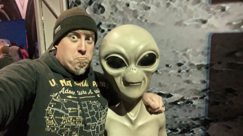 A person in a beanie poses with a life-sized alien figure, making a funny face in a themed setting.