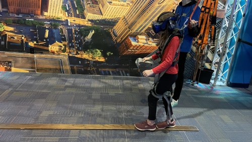 A child wearing a VR headset walks on a narrow beam, guided by an adult in a tech exhibit.