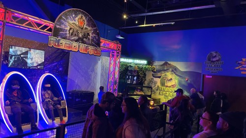 A vibrant arcade featuring "Robo Wars" and "Jurassic Escape," with people waiting in line and interactive gaming stations.