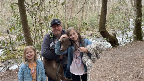 A man holds a dog while two girls smile beside him in a wooded area near a stream.
