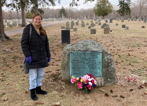 A woman stands beside a gravestone in a cemetery, adorned with flowers, on a cloudy day.