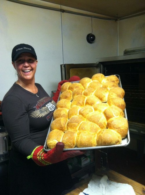 A smiling woman in an apron holds a tray of freshly baked rolls, wearing oven mitts in a kitchen setting.