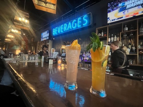 Two colorful cocktails sit on a bar, with a neon "BEVERAGES" sign glowing in the background.
