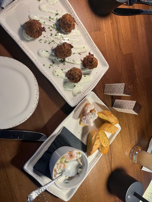 Two plates of food on a wooden table: one with crab cakes and sauce, the other with seafood salad and crackers.