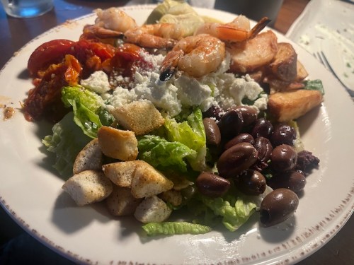 A colorful plate featuring shrimp, olives, lettuce, tomatoes, feta cheese, and crispy bread cubes.