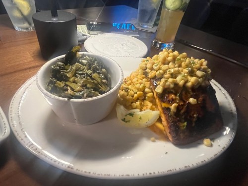 A plate featuring grilled fish topped with corn, served with a side of collard greens and lemon wedges.