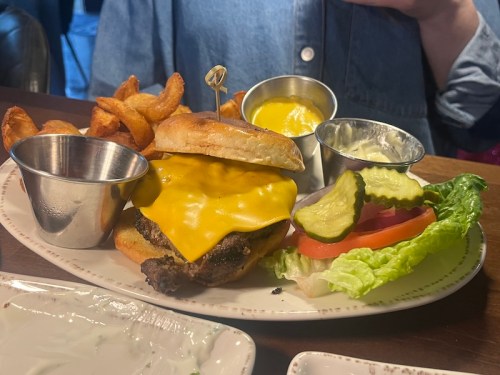 A cheeseburger with melted cheese, served with curly fries, pickles, lettuce, tomato, and dipping sauces on the side.