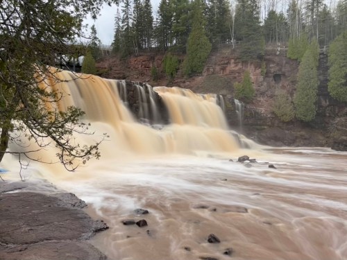 A cascading waterfall flows over rocky terrain, surrounded by lush green trees and a cloudy sky.