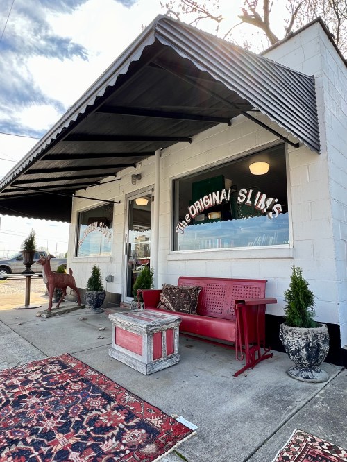 Exterior of a restaurant called "The Original Slim's," featuring a red bench, decorative plants, and a vintage dog statue.