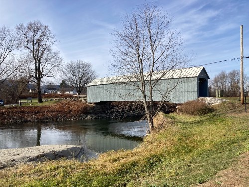 A blue covered bridge spans a calm river, surrounded by bare trees and grassy banks under a clear sky.