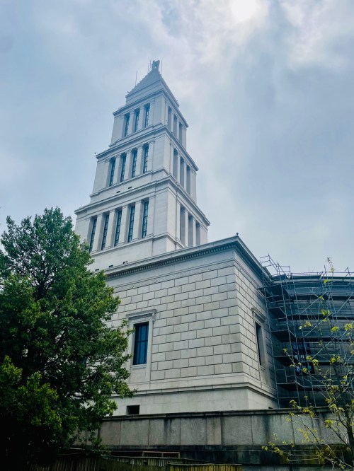 A tall, white stone building with a tower, partially covered in scaffolding, surrounded by trees under a cloudy sky.