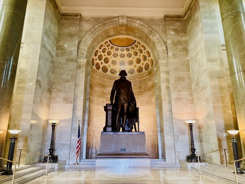 Statue of a historical figure in a grand hall with decorative ceiling and soft lighting, flanked by American flags.