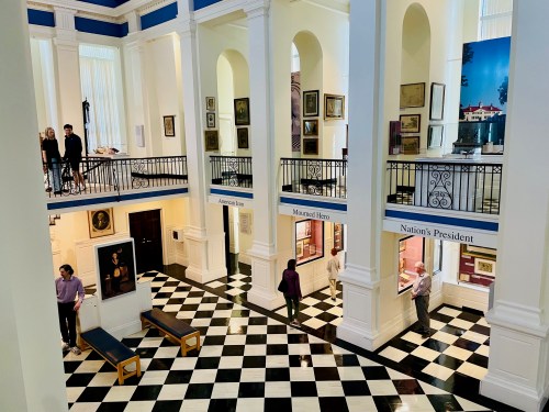Interior view of a museum with a checkered floor, featuring art displays and visitors exploring the exhibits.