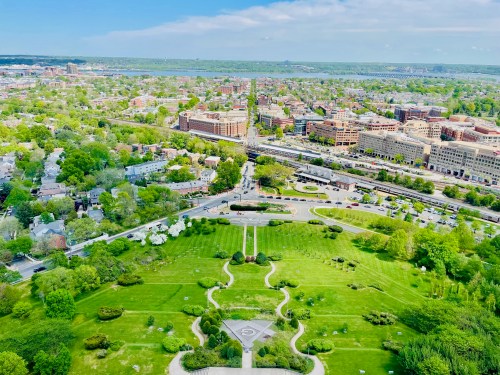 Aerial view of a green park with pathways, surrounded by a cityscape and distant hills under a clear blue sky.