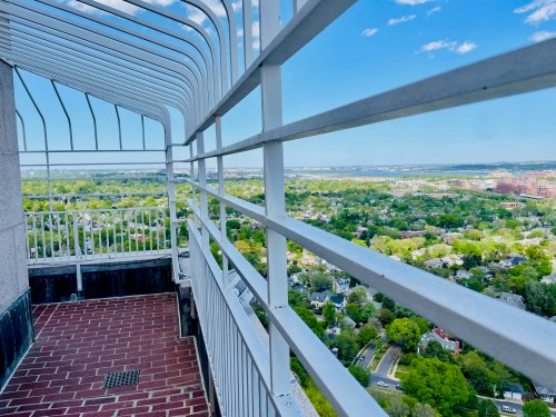 A view from a high balcony with white railings overlooking a green landscape and blue sky.