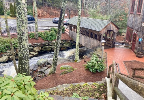 A wooden bridge crosses a stream surrounded by trees, with a parking area and building in the background.