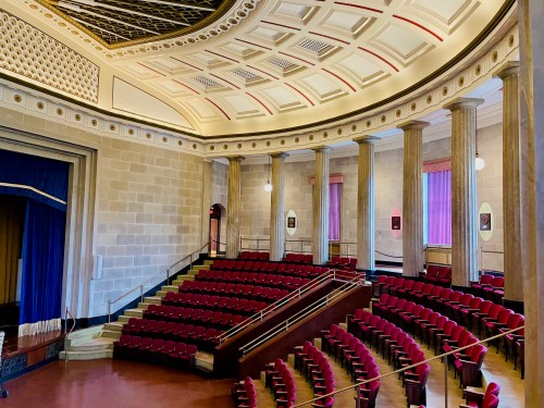 Interior of a theater featuring red seating, tall columns, and a decorative ceiling. Stage area visible in the background.