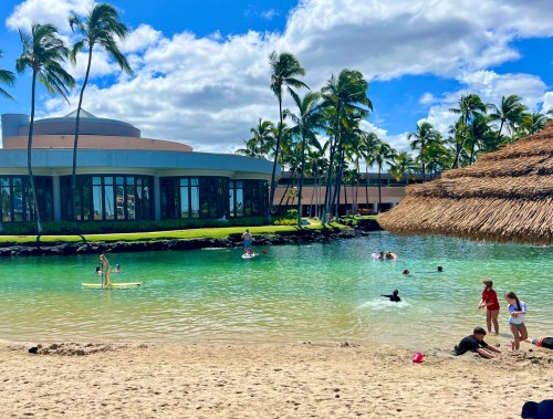 A sunny beach scene with people swimming and paddleboarding near a tropical resort surrounded by palm trees.