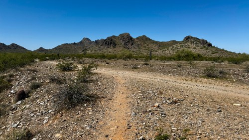 A dirt path winds through a rocky desert landscape with distant mountains under a clear blue sky.