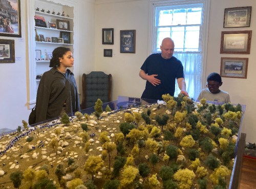 A man explains a model landscape to two children in a room filled with framed pictures and a cozy chair.