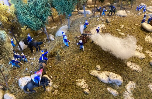 A miniature scene depicting soldiers in blue uniforms, horses, and a cannon in a forested landscape.