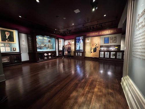 A museum exhibit featuring displays about the Sazerac cocktail, with wooden floors and framed portraits on the walls.
