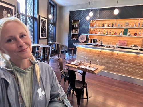 A smiling person stands in a stylish bar with wooden shelves and a variety of bottles in a well-lit space.