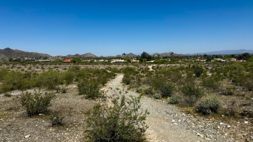 A dry, desert landscape with sparse vegetation and distant houses under a clear blue sky.