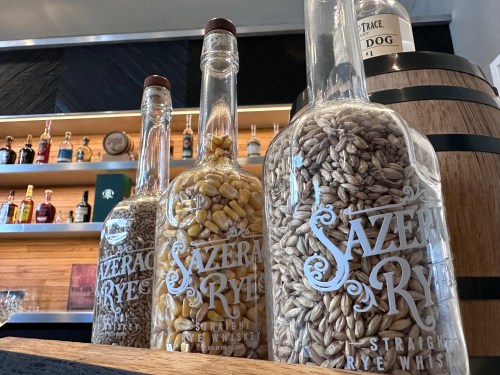 Three glass bottles labeled "Sazerac Rye" filled with grains, displayed on a bar shelf with whiskey bottles in the background.