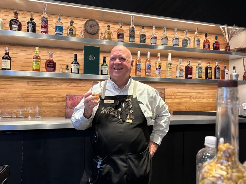 A smiling bartender holds a glass of whiskey, standing in front of a shelf filled with various liquor bottles.