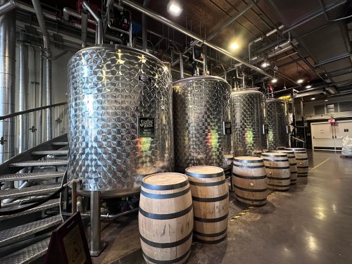 A row of shiny silver fermentation tanks beside wooden barrels in a modern brewery setting.