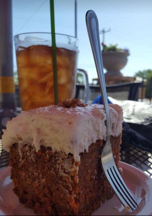 A slice of carrot cake with cream cheese frosting on a plate, next to a glass of iced tea with a straw.